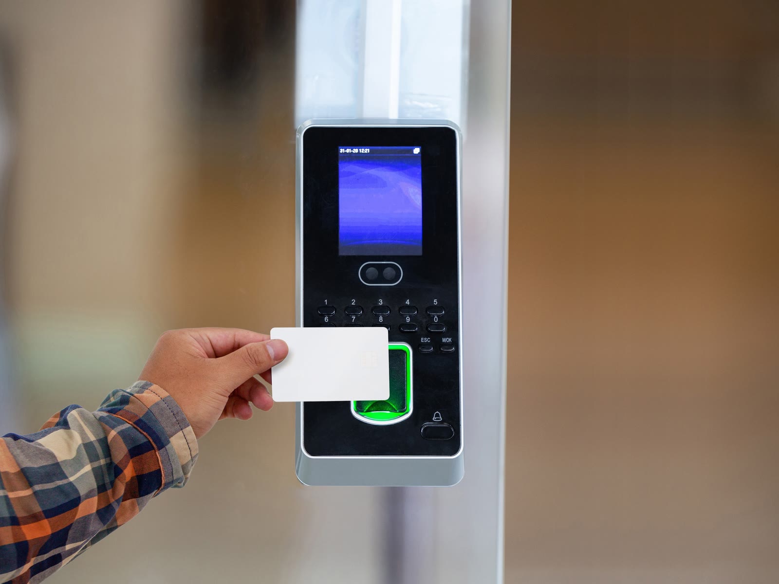 A keycard held to a wall-mounted access control reader with a keypad and fingerprint pad beside a building entrance