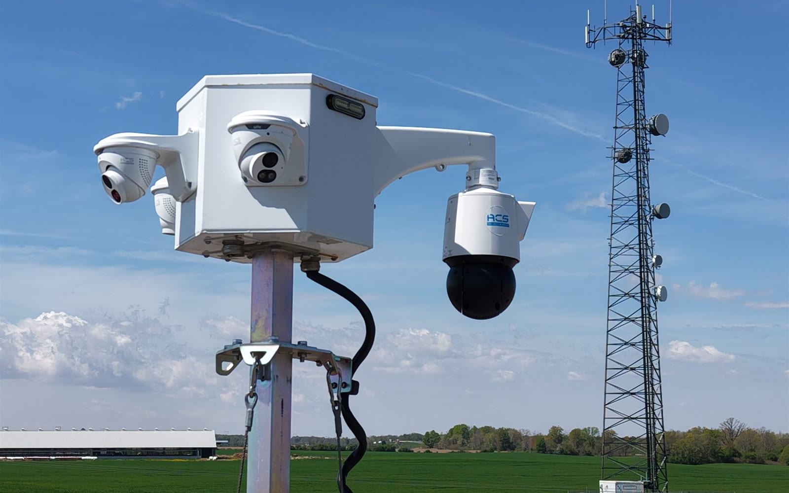 An ACS camera head on a mast with open fields and a communications tower behind