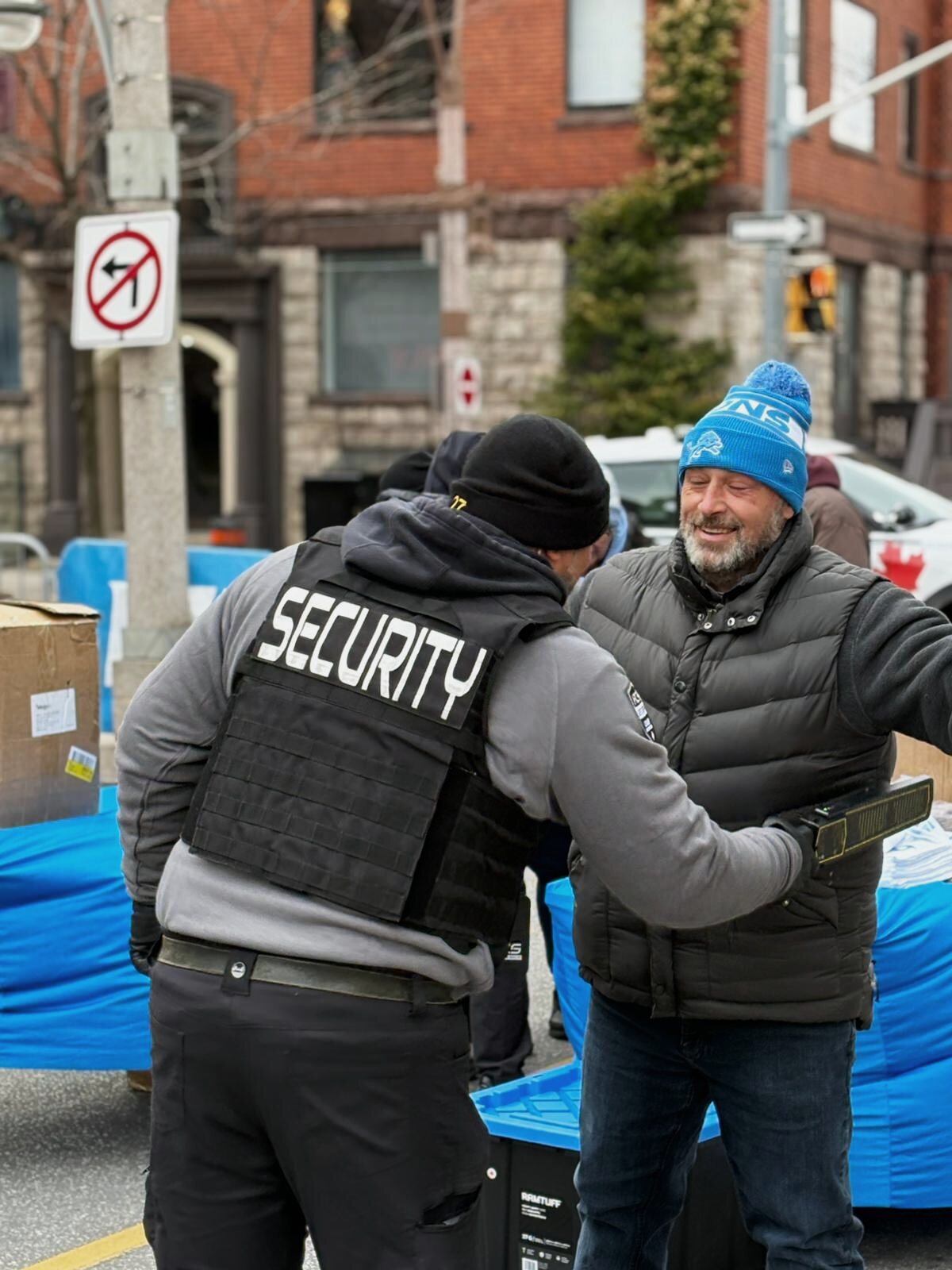 ACS security guard, seen from behind, checking a smiling man with a handheld metal detector at an outdoor street event