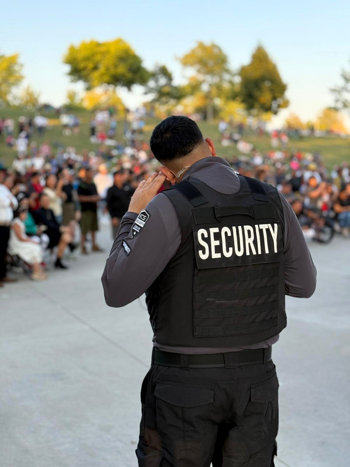 ACS security guard, seen from behind with a hand to his ear, facing a large seated crowd at an outdoor park gathering