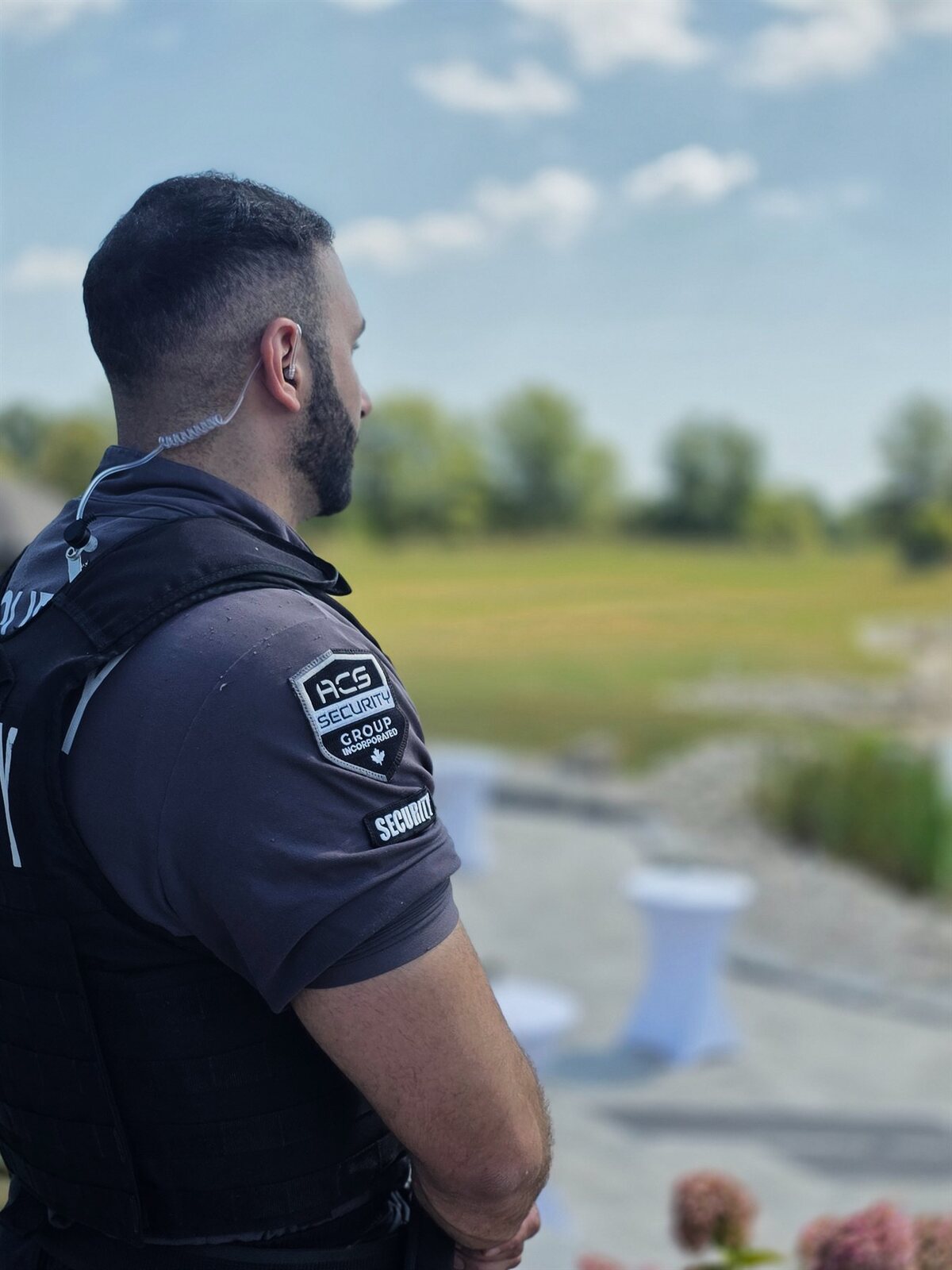 ACS security guard with an earpiece, seen in profile, looking out over an open lawn