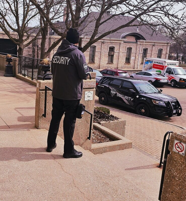 ACS guard in a SECURITY jacket on a sidewalk beside a parked ACS patrol SUV