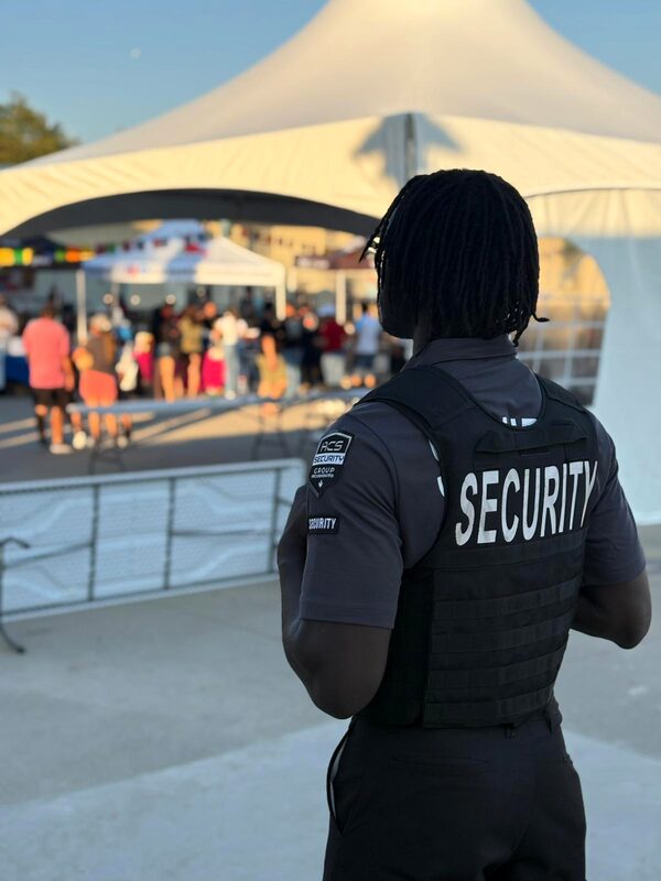 ACS security guard in a security vest watching a crowd gathered under a tent outdoors