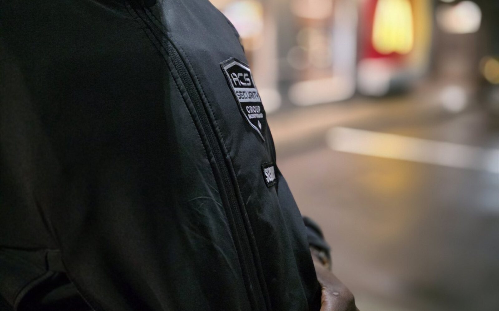 ACS security guard with shoulder patch on night detail outside a lit storefront