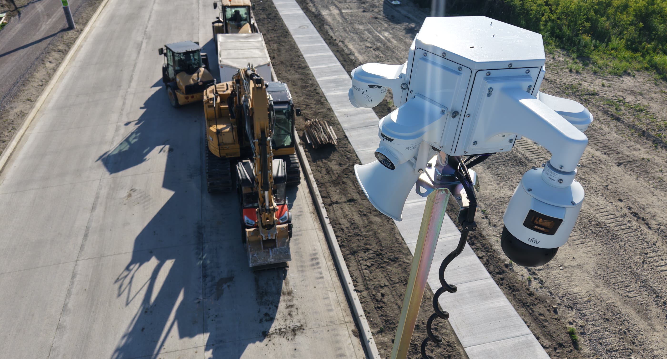 An ACS camera cluster on a mast overlooking an active road construction site, excavator and roller working behind it