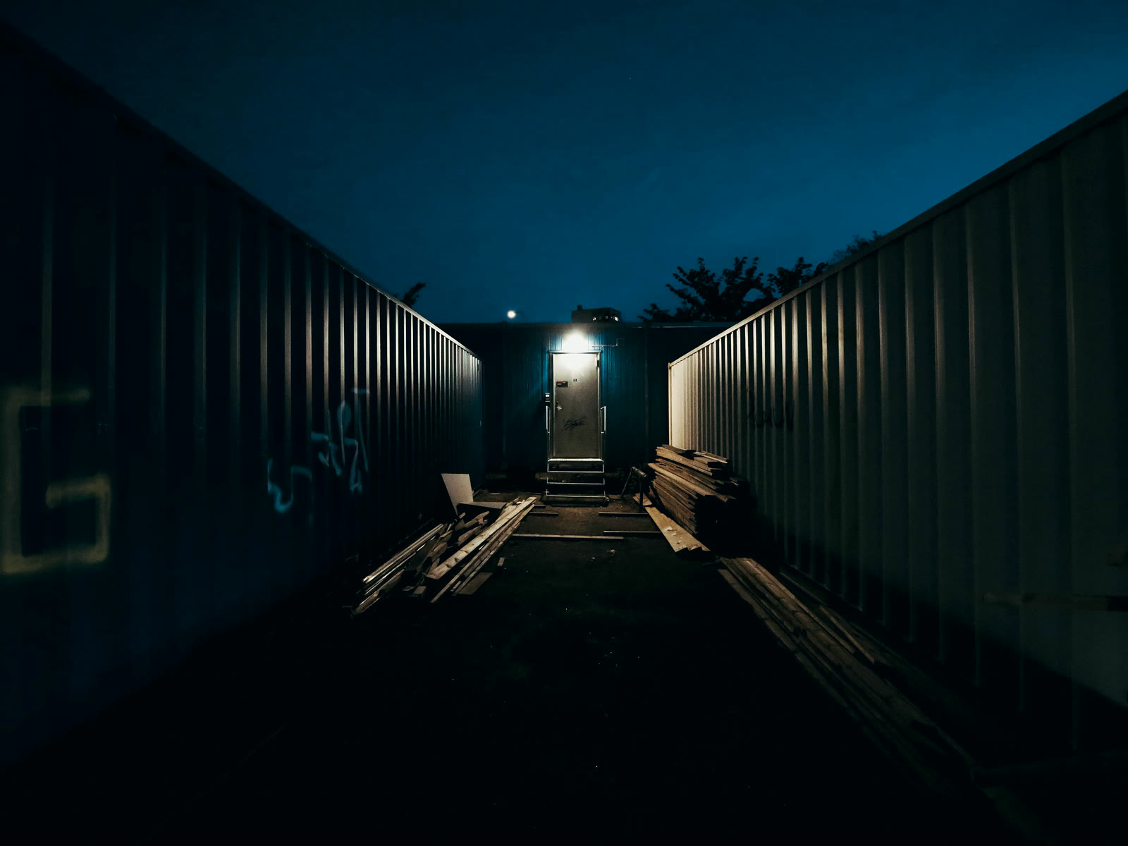 A corridor between two rows of shipping containers at night, one lamp lit at the far end