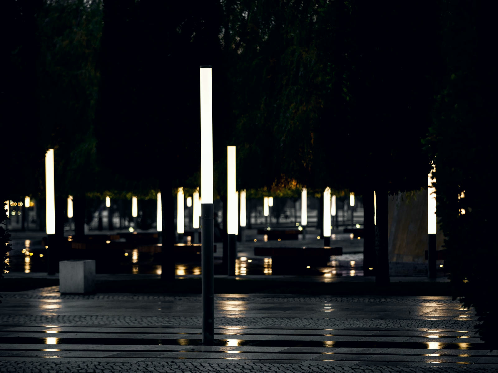 An empty public square at night, tall light columns reflected on wet paving