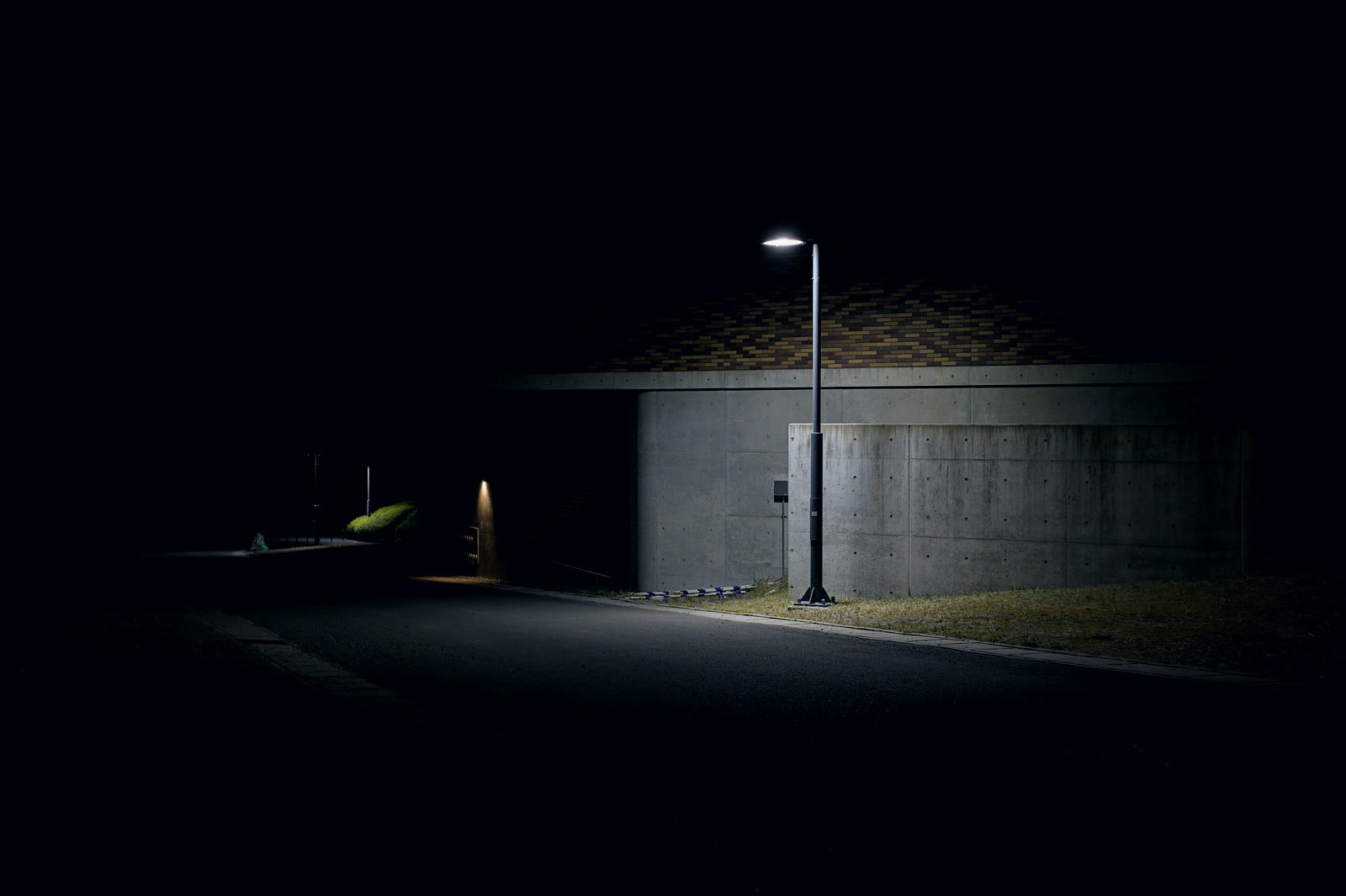 An empty road at night beside a concrete wall, one streetlight lit
